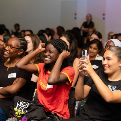A diverse group of young women laugh and cheer in the audience at Next Tech Fest