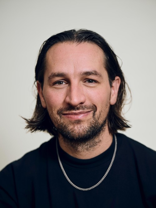 A man with brown hair and wearing a silver chain is looking direclty with a confident expression.