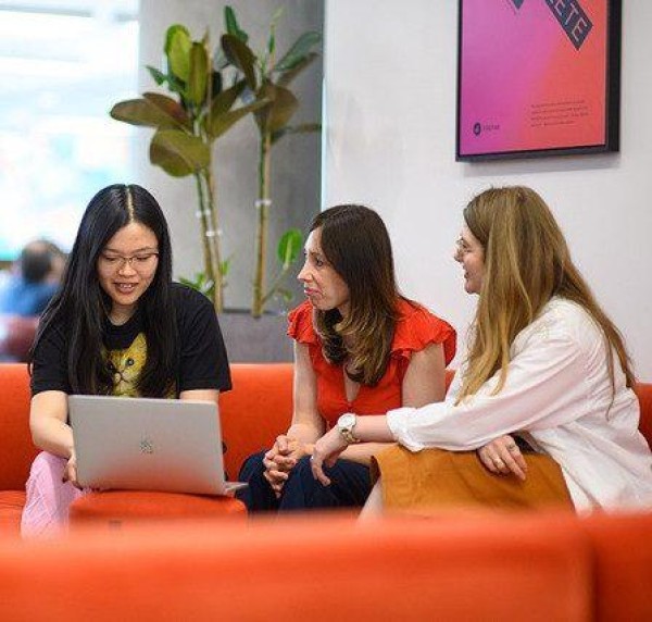 Three women sitting on a red sofa in an office space. One is showing her laptop screen to the others while they look at her intently.