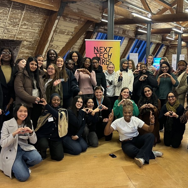 A large group of women standing and kneeling, smiling and posing with their hands making hearts or peace signs in front of a colourful Next Tech Girls banner at the Mastek Insight Day in Manchester.