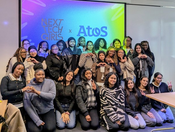 A group of 25 young women smiling, making heart hands or peace signs, with a colourful background showing Next Tech Girls and Atos logos.