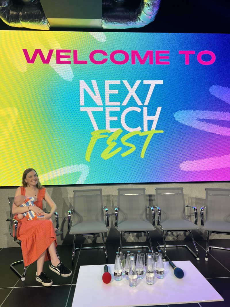 Emily Hall-Strutt of Next Tech Girls holding a baby and sitting in front of a screen that says 'Welcome to Next Tech Fest'.