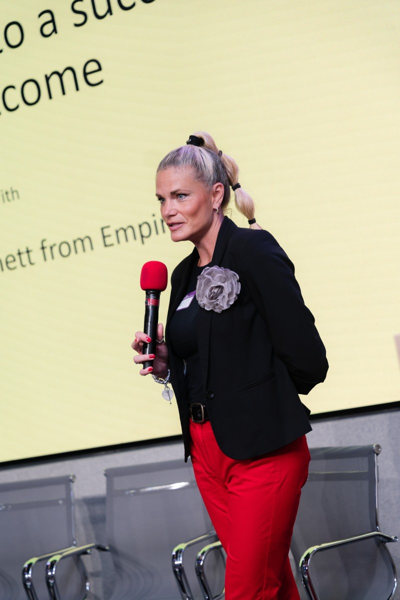 A woman with blonde hair holding a microphone and presenting in front of a large screen.