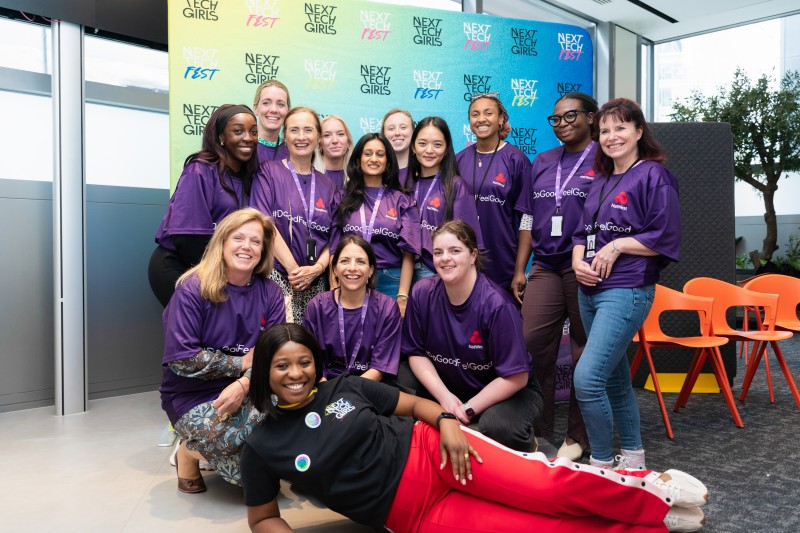 A group of women wearing purple Natwest t-shirts are smiling together for a group picture in front of the Next Tech Girls colourful backdrop. The woman at the front of the group is wearing a Next Tech Girls t-shirt and posing with her hand on her hip, lying sideways on the ground.